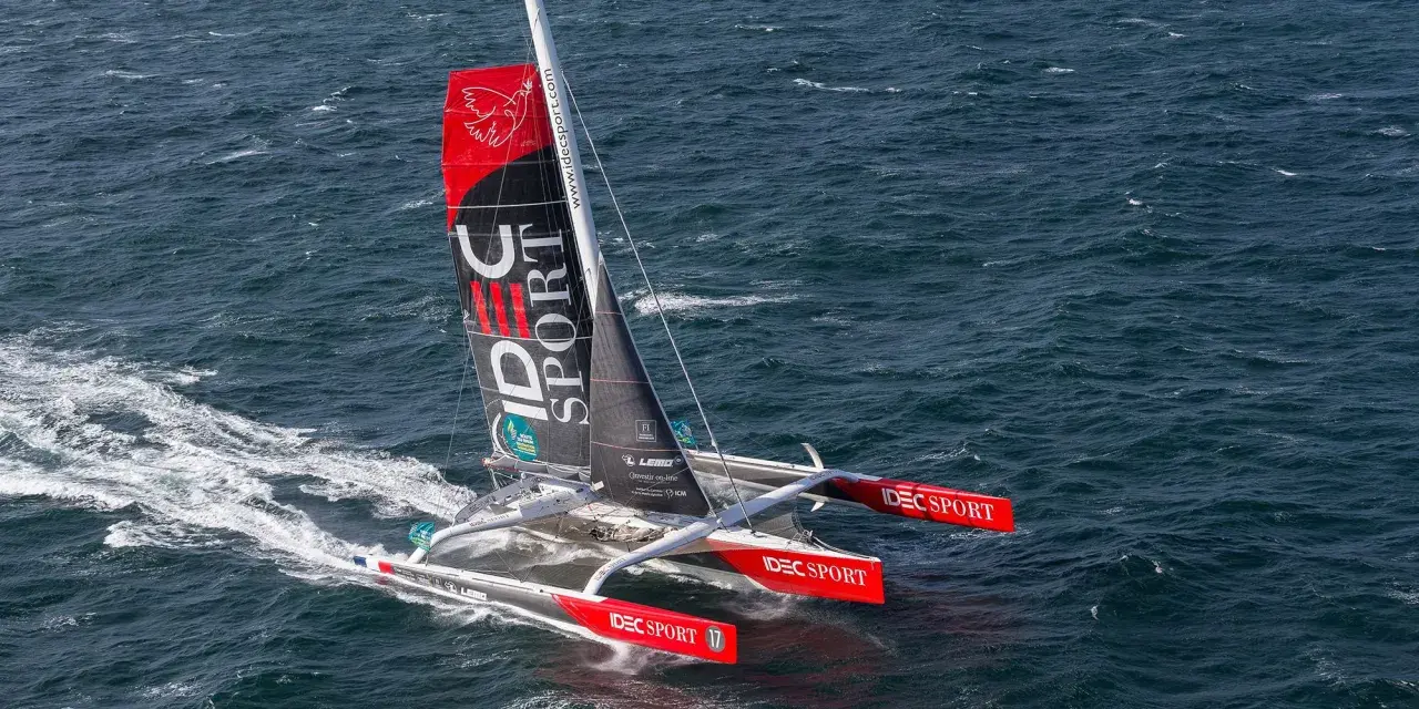 Le trimaran IDEC SPORT fend les vagues, une voile rouge et noire ornée d'un oiseau blanc.