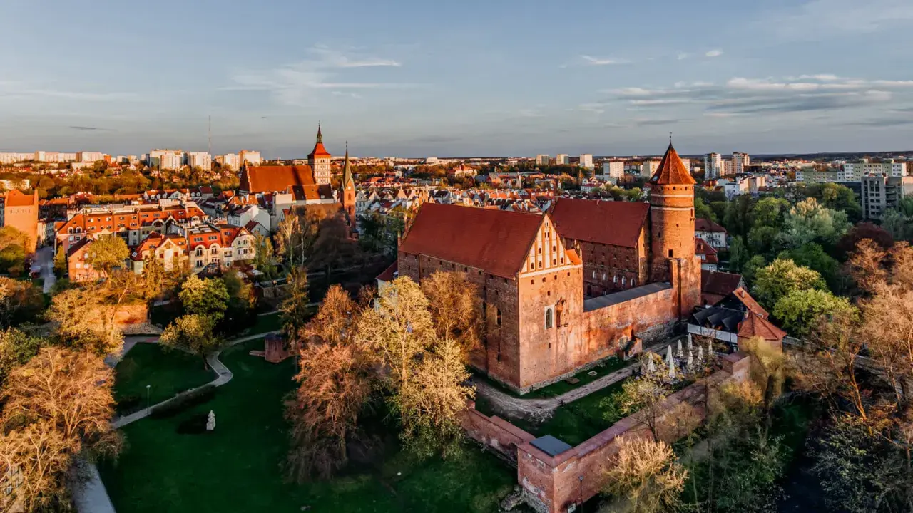 Zachód słońca nad zamkami krzyżackimi na Mazurach. Potężne ceglane mury i wieże w blasku złota, otoczone zielenią i miastem.
