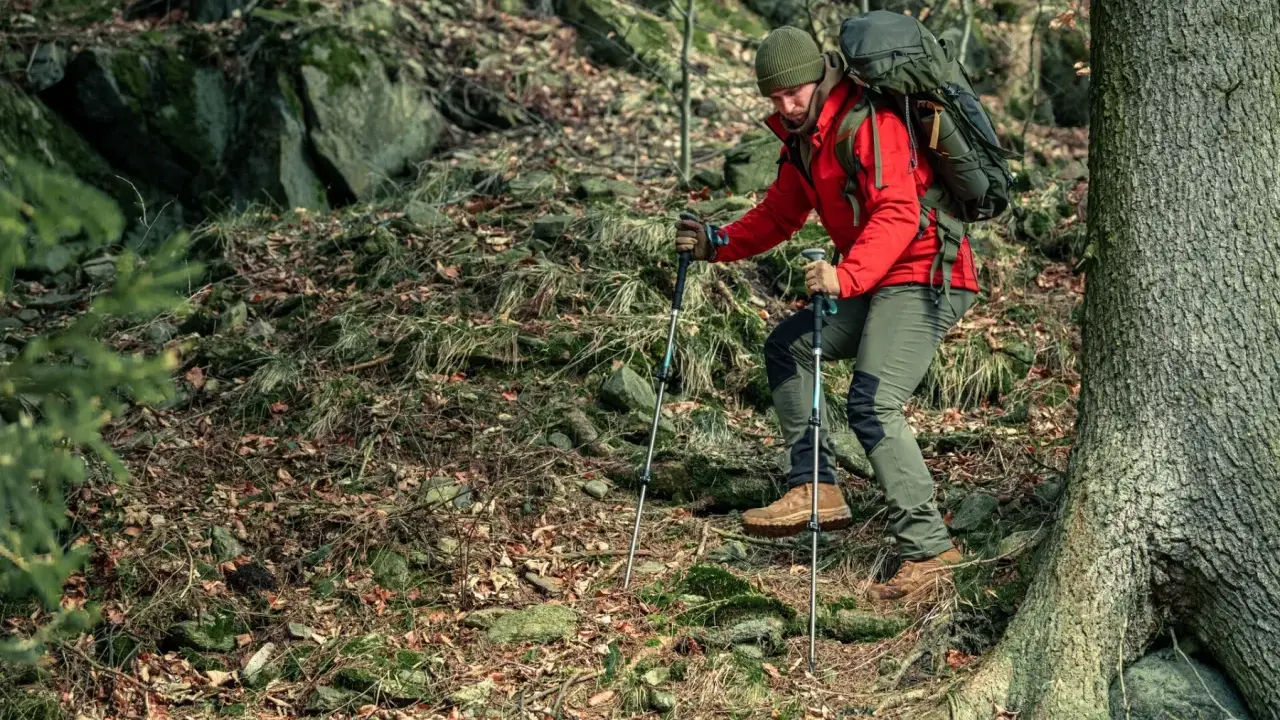 Mężczyzna w czerwonej kurtce i zielonej czapce z plecakiem wspina się po kamienistym, leśnym szlaku, używając kijków. To jest właśnie trekking!