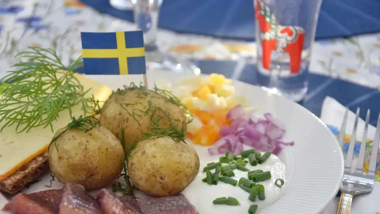 En tallrik med midsommar mat: sill, färskpotatis, ost, ägg och rödlök. En svensk flagga vajar stolt.