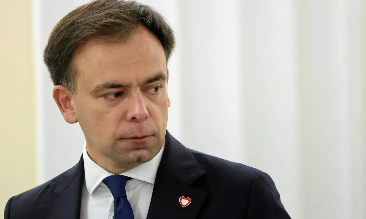 Minister finansów w granatowym garniturze i niebieskim krawacie, z przypinką w kształcie serca na klapie marynarki.