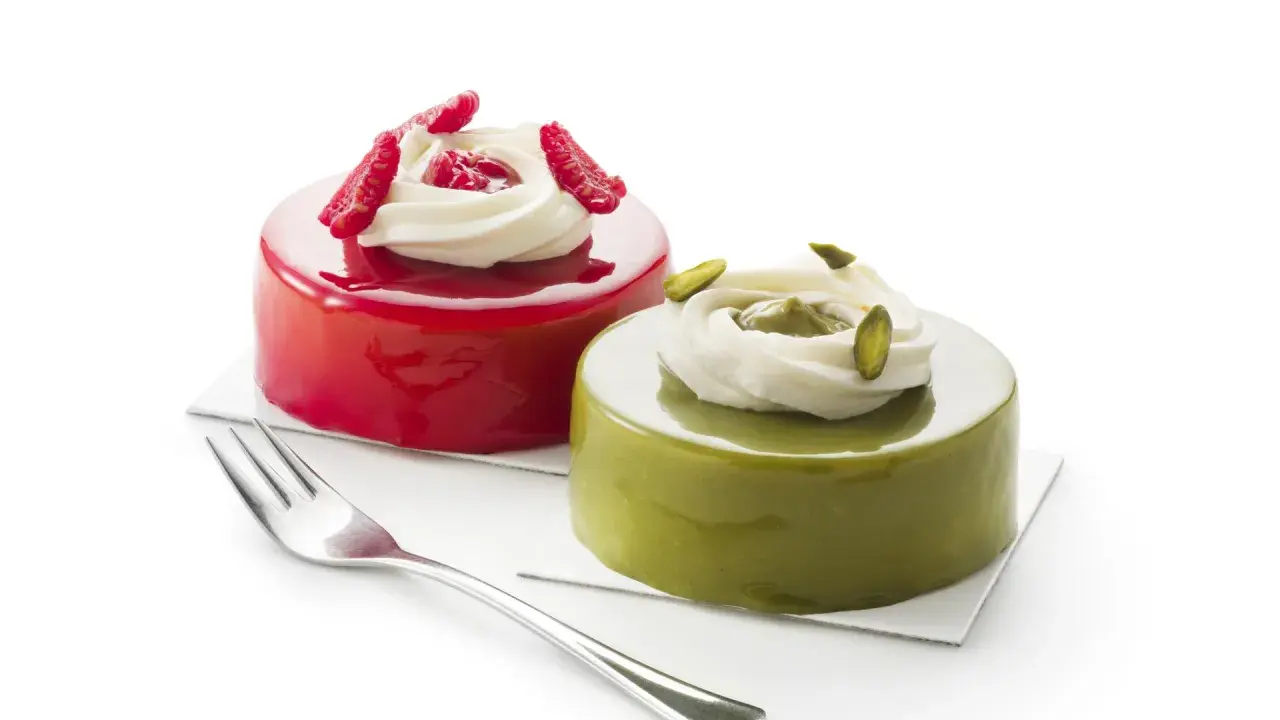 Deux petits gâteaux brillants, l'un rouge vif, l'autre vert avec un glaçage pistache, ornés de crème et de fruits.