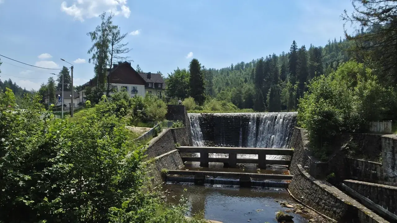 Kamienny stopień wodny na Jeziorze Czerniańskim, z wodą spływającą kaskadami. W tle zielone wzgórza i domy.