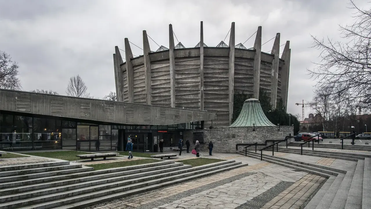 Monumentalny budynek w stylu brutalizm z betonowymi żebrami i szklanym wejściem. Schody prowadzą do wejścia, a wokół rosną drzewa.