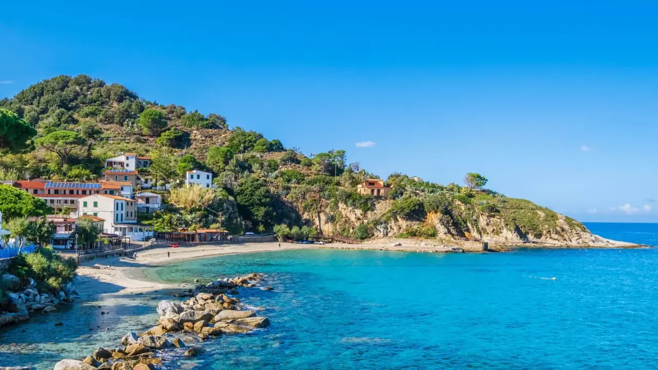 En pittoresk kustby på Elba, Italien, med vita hus som klättrar uppför en grönskande kulle. Klart turkost vatten sköljer mot en sandstrand.