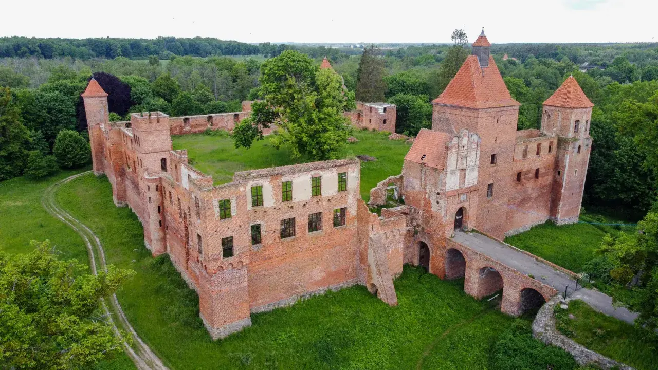 Ruiny ceglanego zamku w Szembarku otoczone zielenią. Widać most nad fosą, wieże i fragmenty murów obronnych.