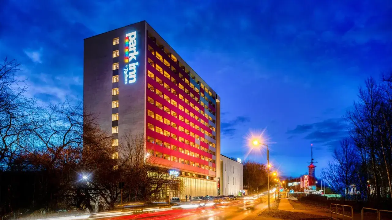 Wieczorem rozświetlony hotel Park Inn w Katowicach z kolorowym logo. Na ulicy widać światła samochodów.