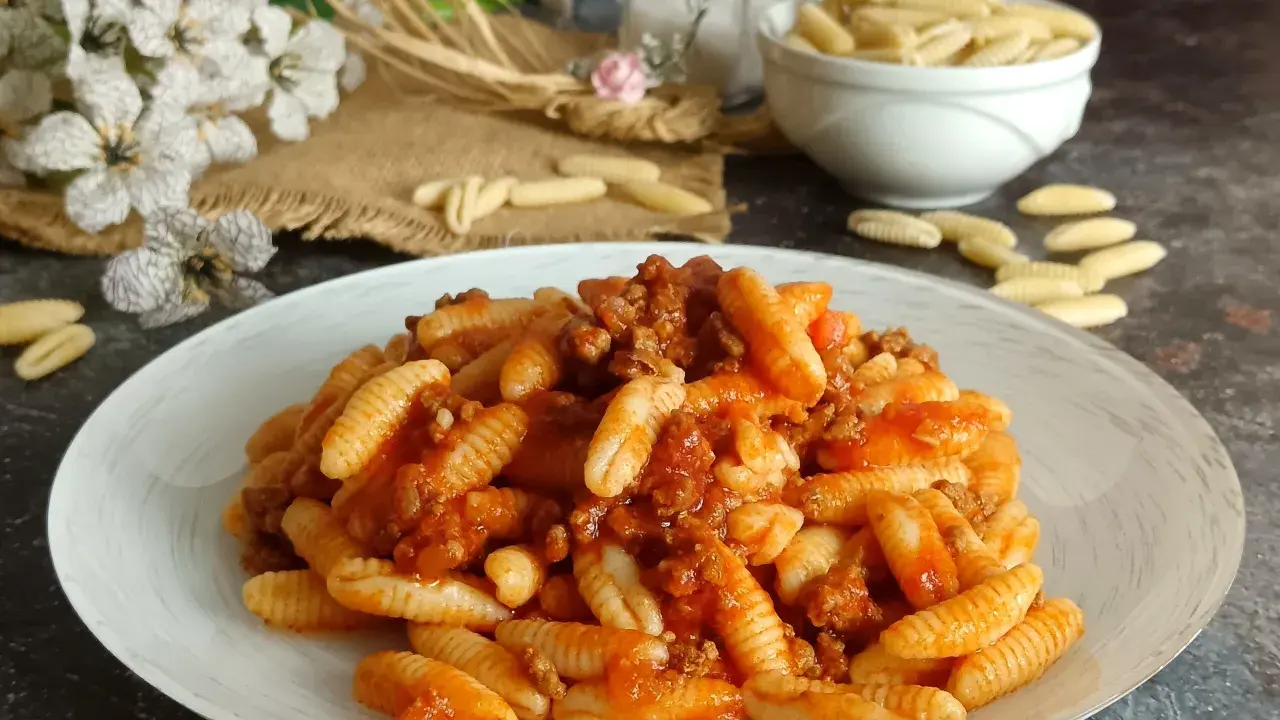 Un piatto invitante di malloreddus alla campidanese, conditi con un ricco ragù di carne. Sullo sfondo, fiori bianchi e un ciotola di pasta cruda.