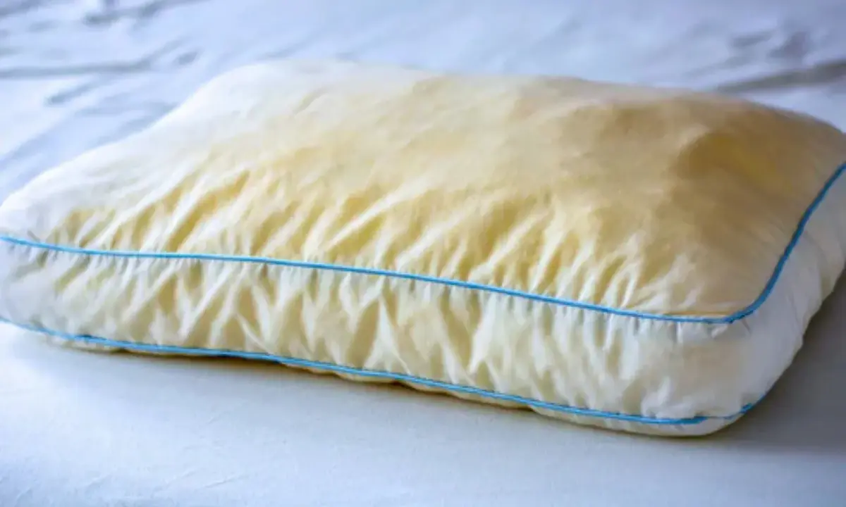 Ein vergilbtes Kissen mit blauen Nähten, das auf einem weißen Bett liegt. Gelbe Flecken deuten auf möglichen Schimmel hin.