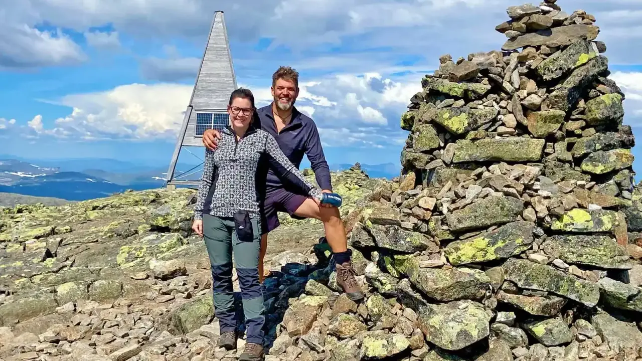 Två glada vandrare njuter av utsikten från en bergstopp under en vandring i Sverige. En stenröse och en modern träkonstruktion med solpaneler syns i bakgrunden.