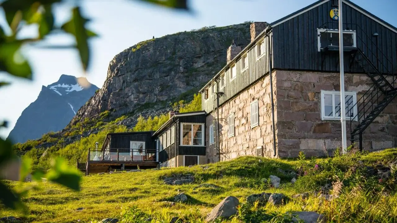 STF Kebnekaise fjällstation med stenfasad och mörk träpanel, belägen vid foten av ett berg. Grönskande sluttning i förgrunden.
