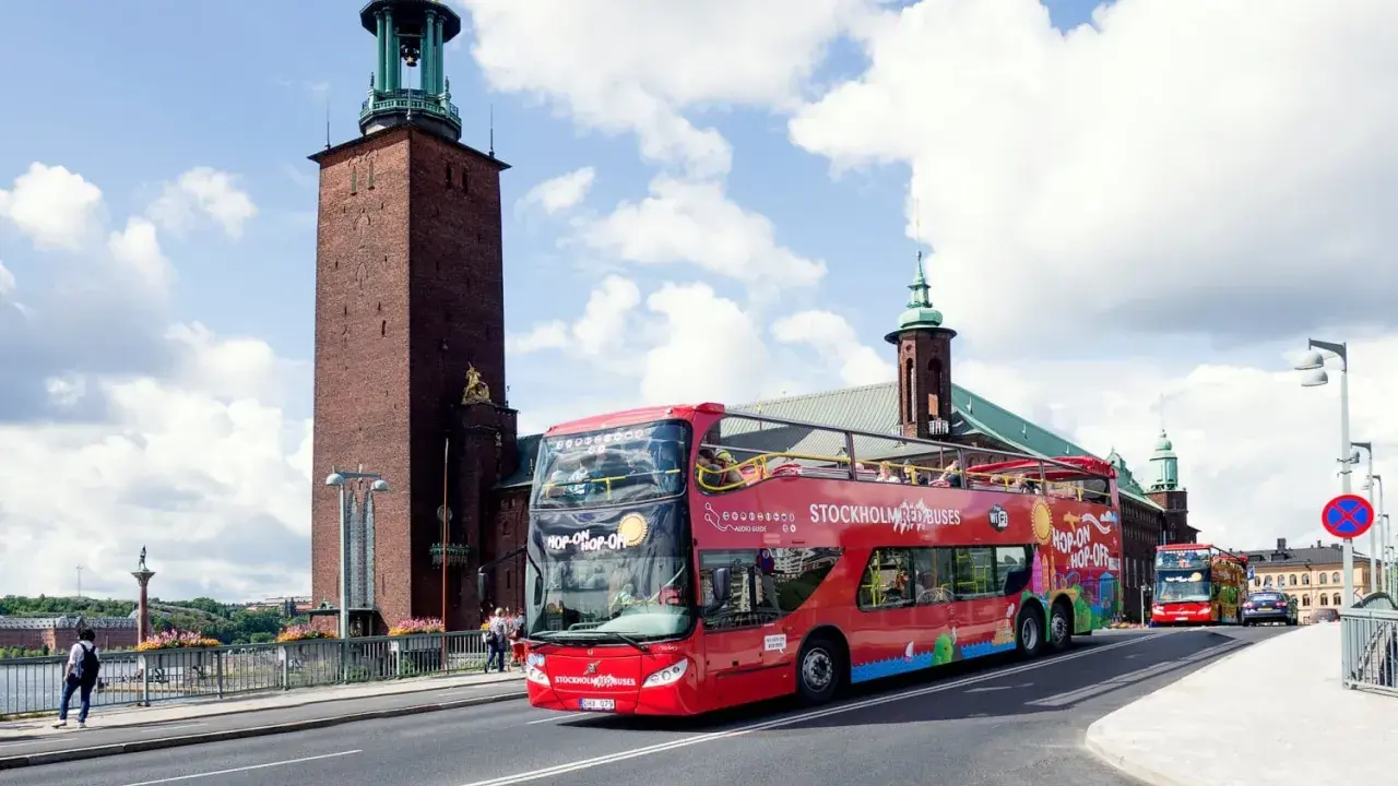 Röd dubbeldäckare med "Hop-on Hop-off Stockholm Buses" på väg förbi Stadshuset. Perfekt för stockholm turistinformation.
