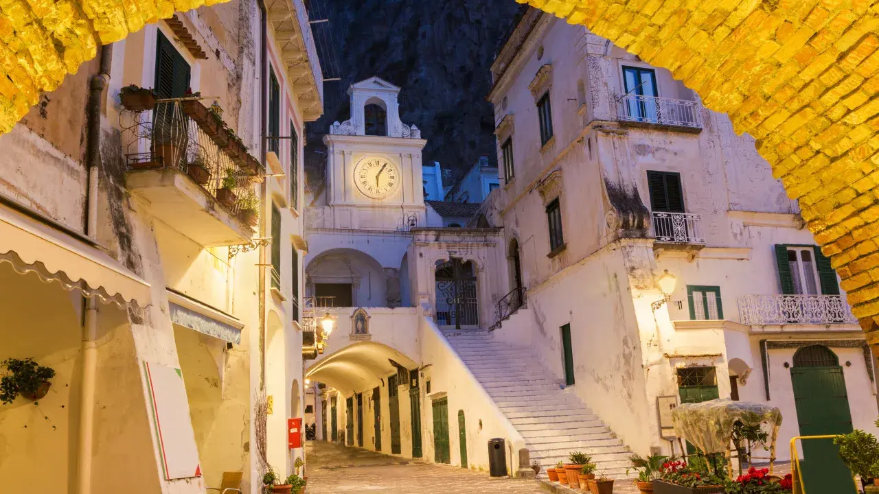 Un vicolo illuminato ad Atrani, con un arco giallo in primo piano e un campanile con orologio sullo sfondo.