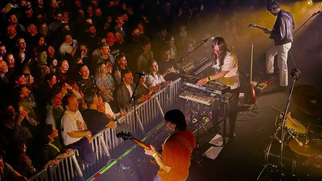 Publik njuter av Altin Gün i Stockholm. En gitarrist spelar, en keyboardist bakom Roland-syntar, och en entusiastisk publik bakom stängsel.