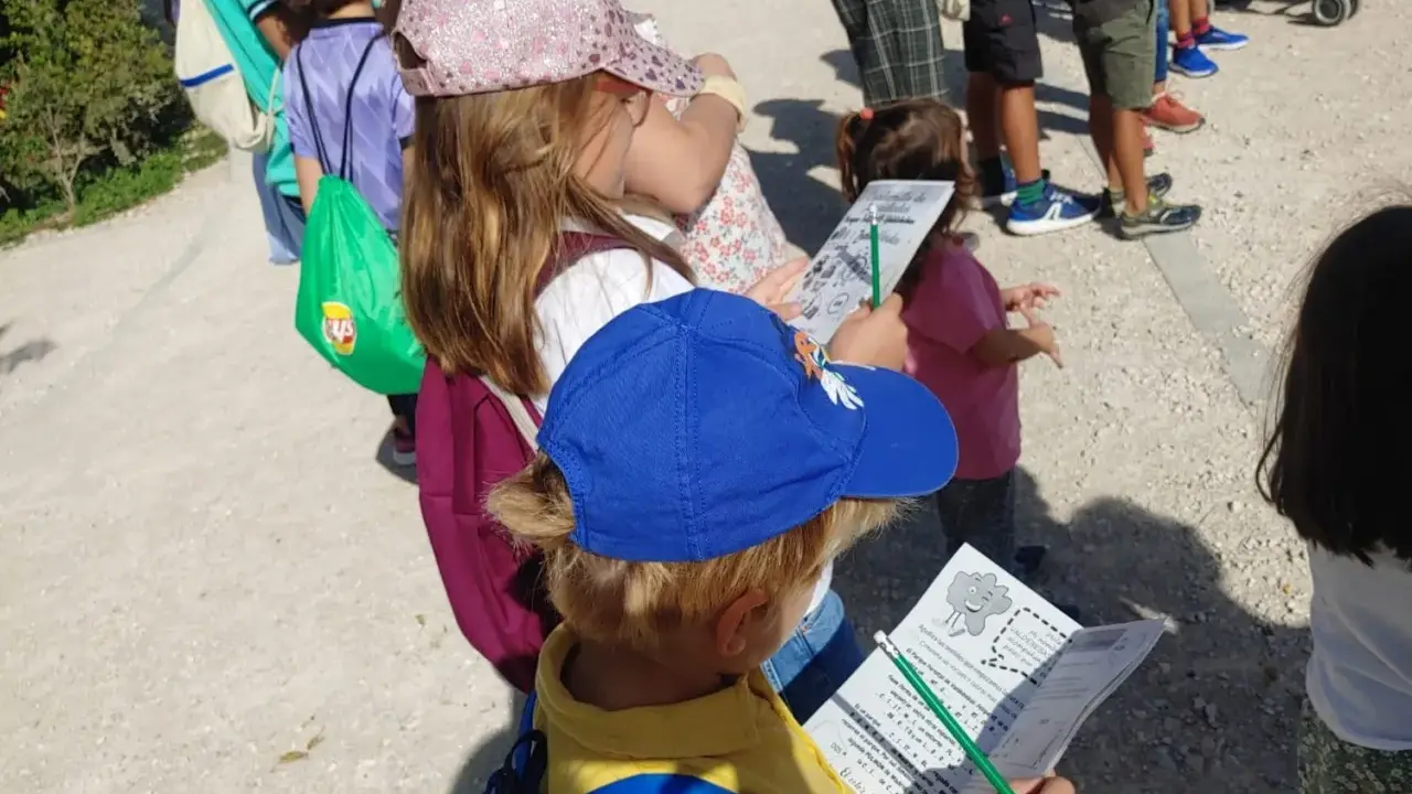 Niños con gorras y mochilas, algunos con folletos, explorando el **parque de Valdebebas** en un día soleado.