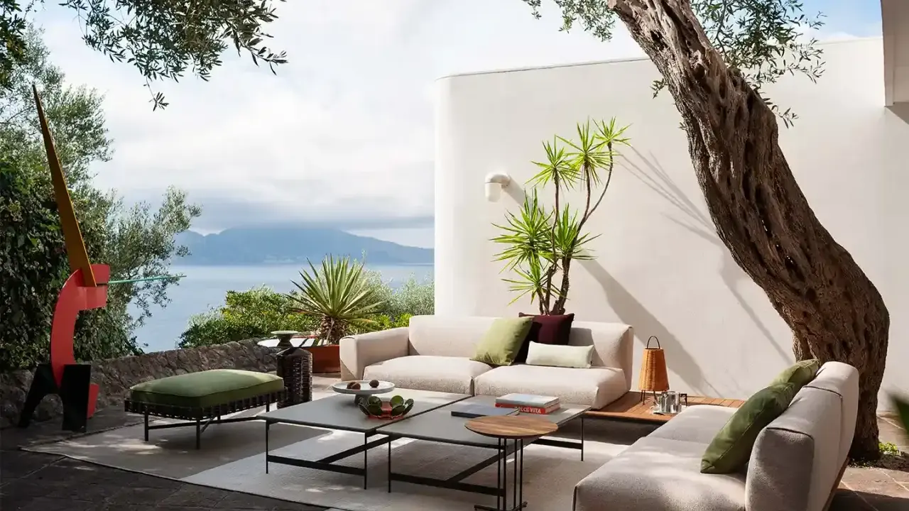 Arredare un terrazzo scoperto con divani chiari, tavolini bassi e un pouf verde, con vista mare e un ulivo secolare.