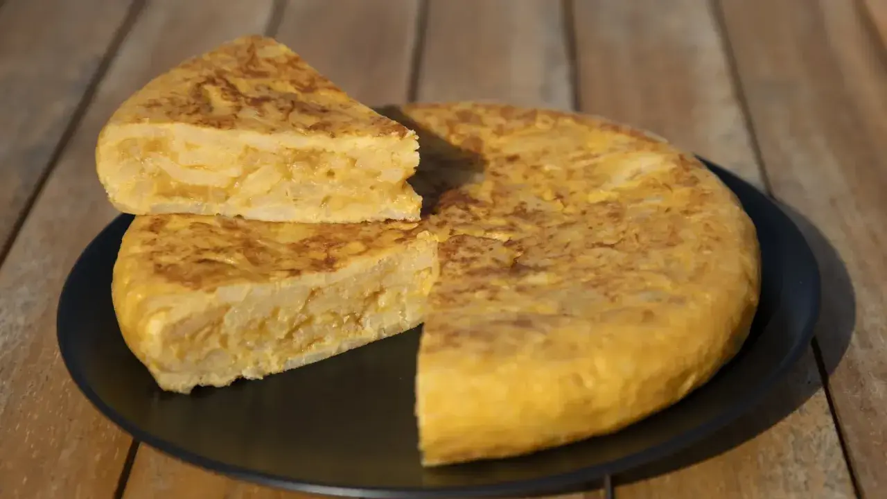 Una deliciosa tortilla de patatas, con una porción cortada y apilada, servida en un plato negro sobre una mesa de madera.