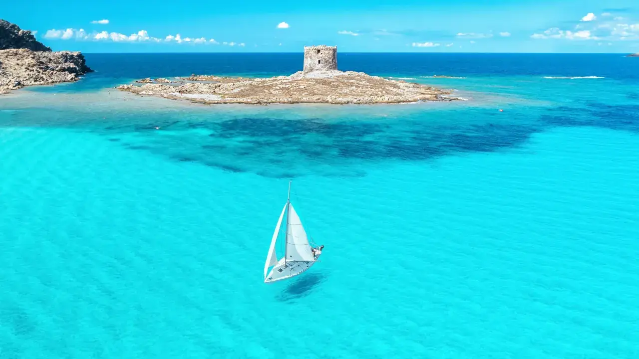 En segelbåt glider fram på turkost vatten mot en gammal vakttorn på en ö. Perfekt för din resa till Sardinien tips!