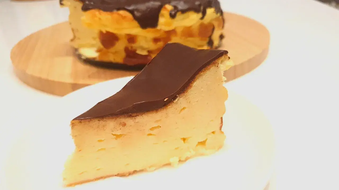 Kawałek puszystego keto sernika z mascarpone, polany czekoladą. Cały sernik w tle.