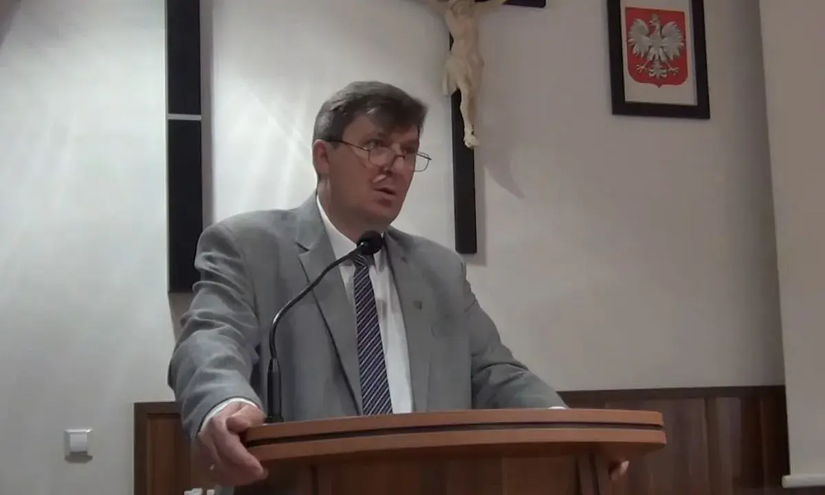 Mężczyzna w garniturze przemawia, za nim krzyż i godło Polski. Może mówi o cnotach kardynalnych.
