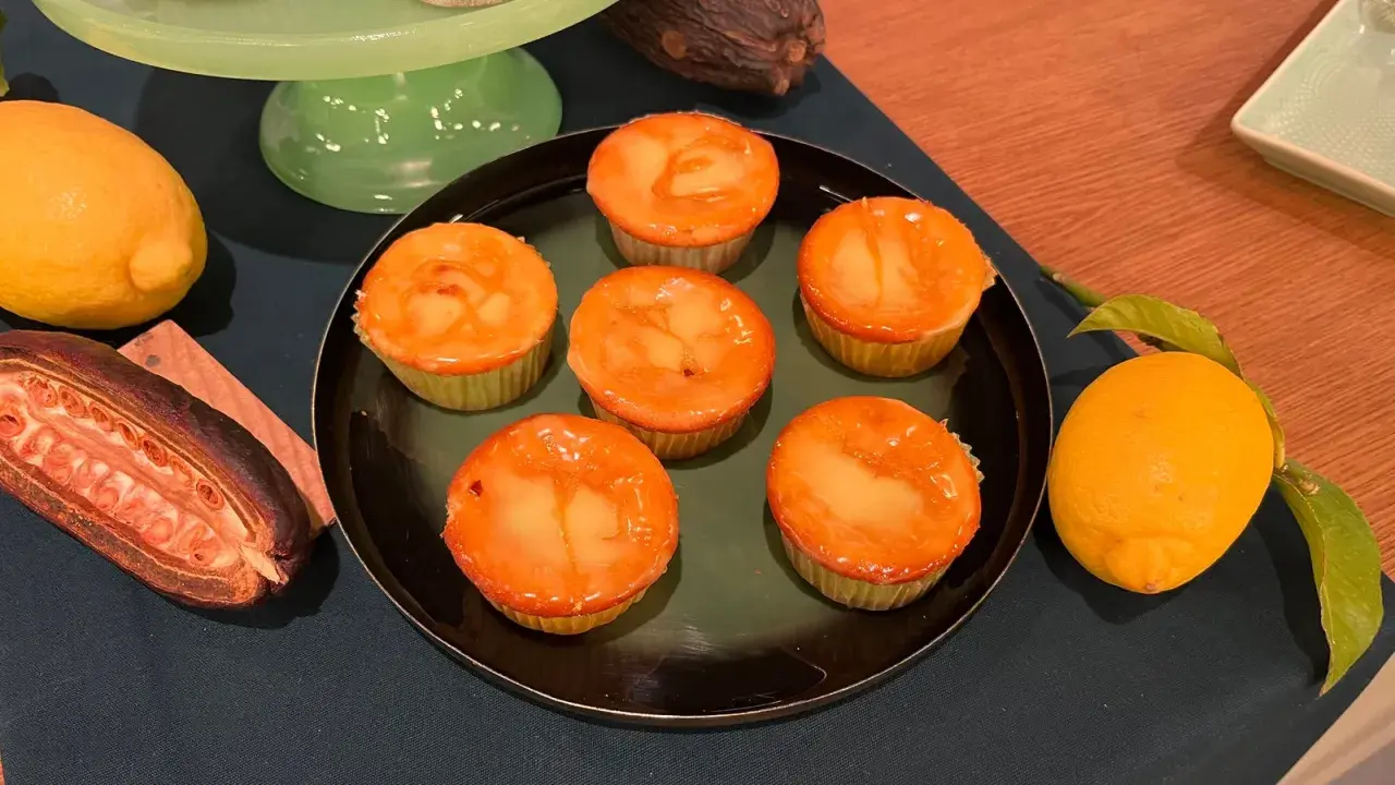 Sechs Zitronenmuffins mit glänzender Glasur auf einem Teller, daneben frische Zitronen und eine Kakaofrucht. Ein leckeres zitronenmuffins rezept!