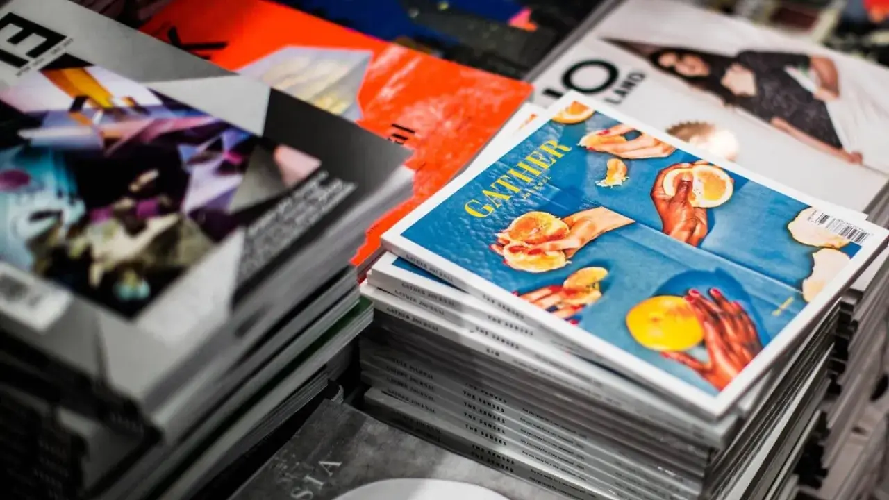 Stosy magazynów, jeden z nich z okładką "GATHER" i pomarańczami, gdzie każdy detal, od tekstury owoców po odbicia światła, podkreśla piękno koloru spotowego.