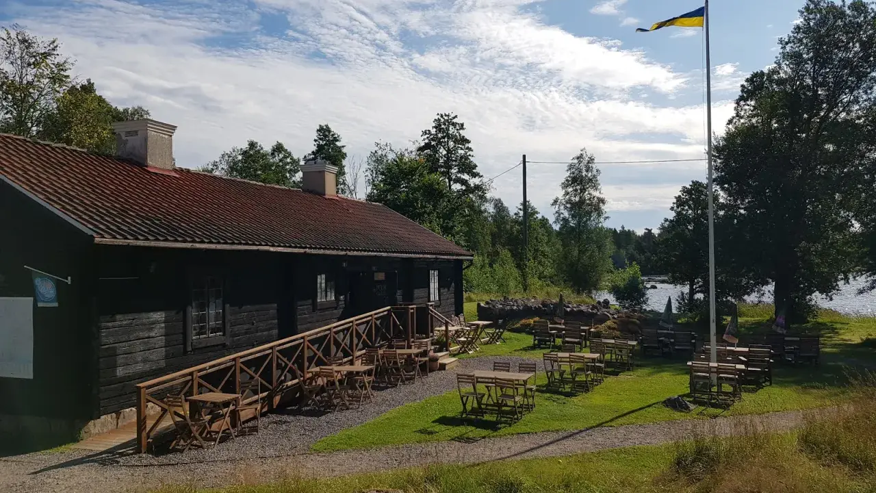 Mysigt tiveden boende med uteservering vid vattnet. En svensk flagga vajar i vinden.