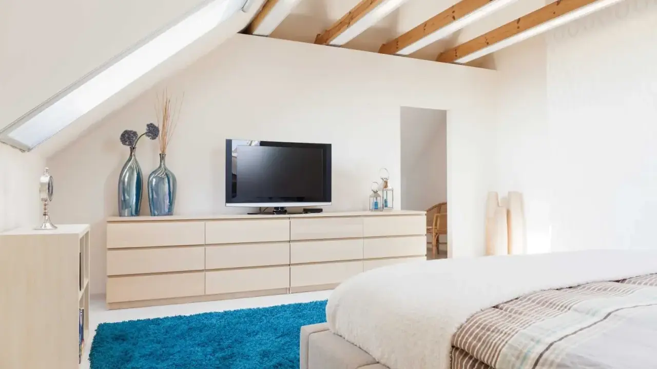 Schlafzimmer im Dachgeschoss, ideal zum Dachboden ausbauen. Helle Farben, Holzbalken und ein blauer Teppich schaffen Gemütlichkeit.
