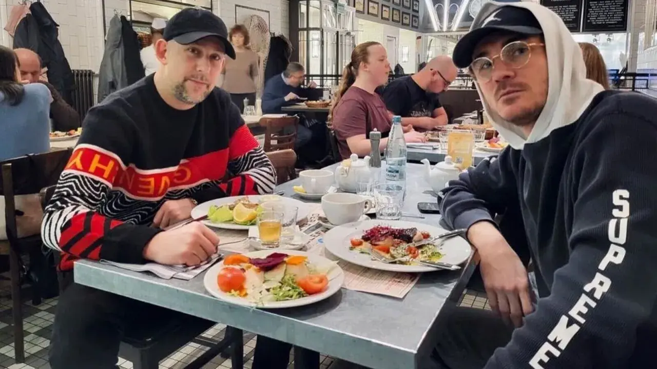 Dwóch mężczyzn, jeden w bluzie z napisem "Givenchy", drugi w bluzie "Supreme", siedzi przy stole w restauracji. Wyglądają jak bary raperzy, którzy wybrali się na wspólny posiłek.