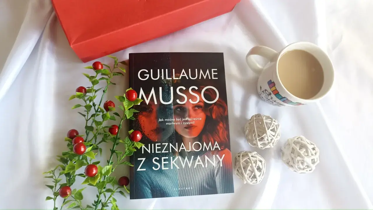 Książka "Nieznajoma z Sekwany" Guillaume'a Musso leży obok kubka kawy i czerwonych ozdobnych kulek.