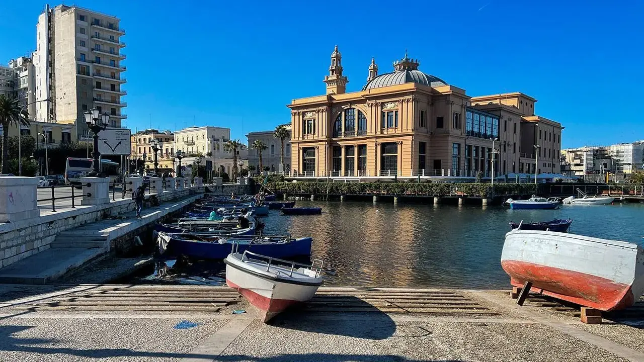 Barche ormeggiate nel porto di Bari, con il Varco della Vittoria e il Teatro Margherita sullo sfondo, sotto un cielo azzurro.