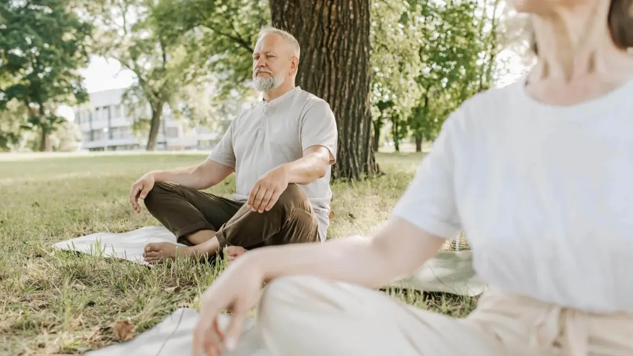 Två personer sitter i lotusställning på gräsmattan under ett träd. De utövar liggande meditation i en lugn parkmiljö.