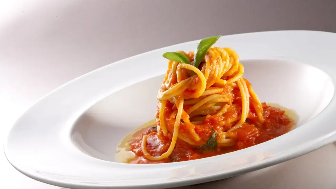 Un nido di spaghetti conditi con una salsa rossa vibrante di pomodorini freschi, guarnito con foglie di basilico verde brillante, servito in un piatto bianco elegante.