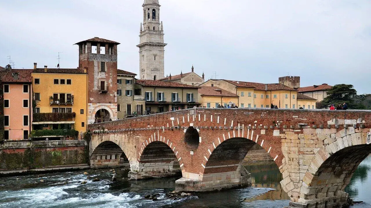 En vacker tegelbro korsar en flod i Verona, Italien. Färgglada hus och ett högt klocktorn syns i bakgrunden.