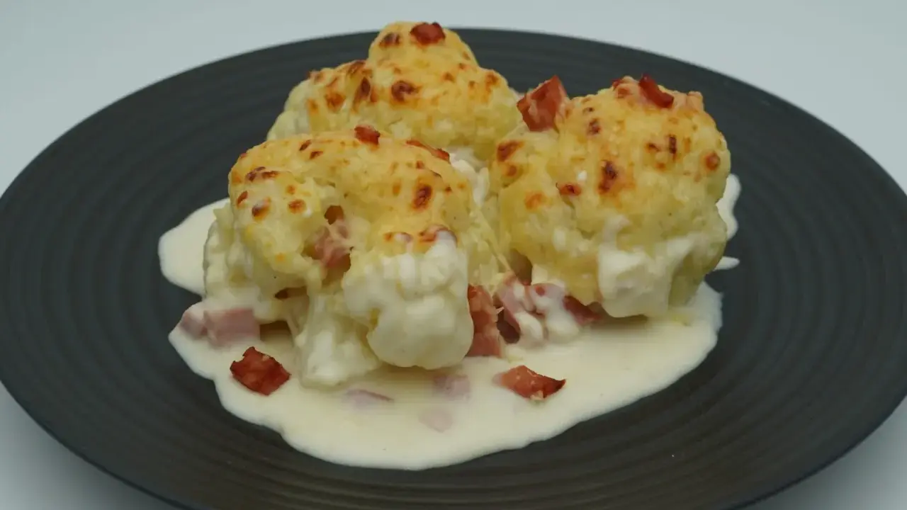 Tres porciones de coliflor gratinada con bechamel y trocitos de jamón, servidas en un plato oscuro.