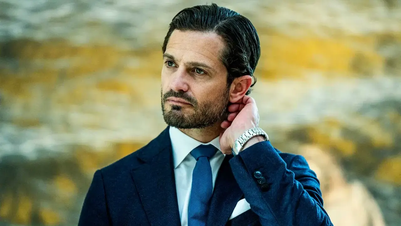 Prins Carl Philip i mörkblå kostym, vit skjorta och blå slips. Hans hand vilar bakom örat, en detalj som speglar hans intresse för carl philip design.