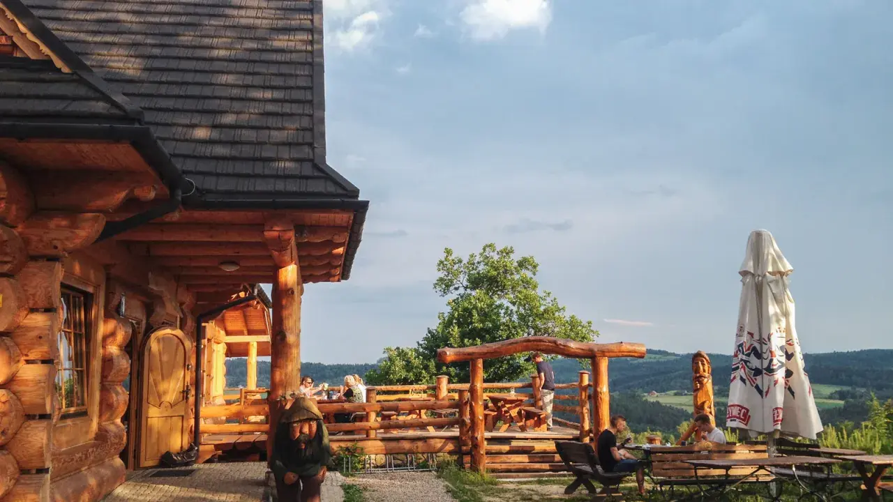 Drewniana bacówka Jamna z tarasem, gdzie goście jedzą, a obok stoi parasol. Menu bacówki Jamna kusi widokiem.