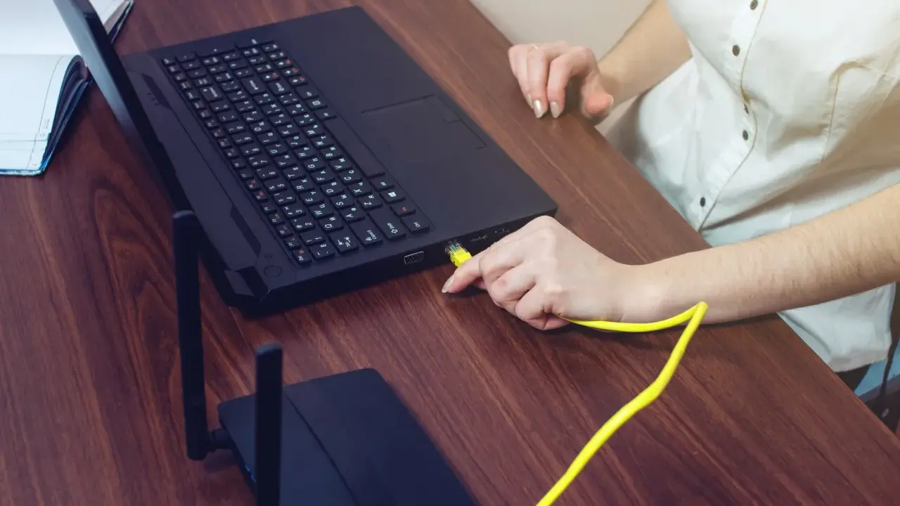 Ręka podłącza żółty kabel do laptopa, pokazując, czy można podłączyć monitor do laptopa. Obok leży router.