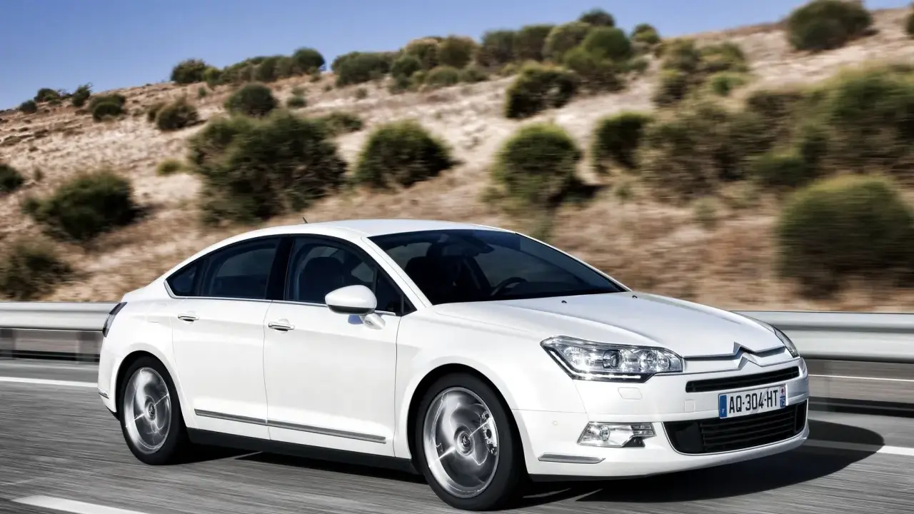 Biały Citroen C5 2.0 HDI pędzi po pustej drodze, otoczony suchymi, pagórkowatymi krajobrazami.