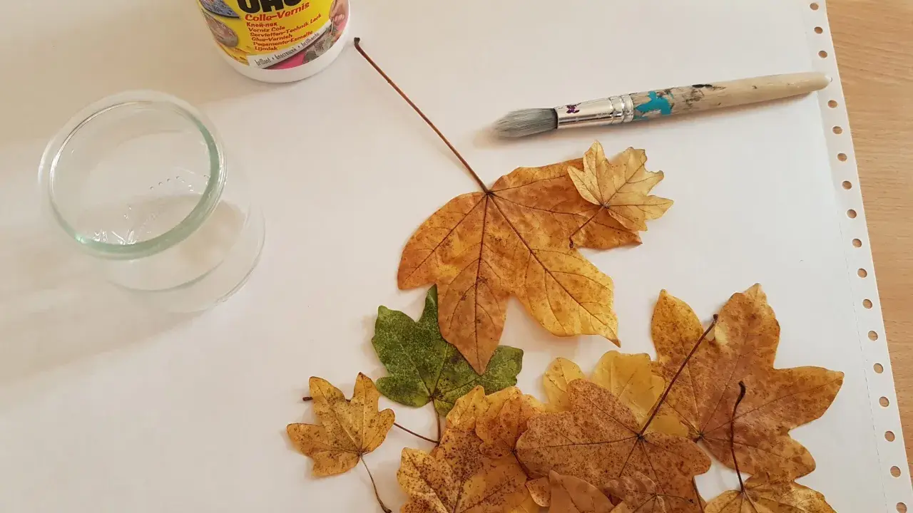 Préparation d'une décoration avec des feuilles d'arbre séchées, de la colle UHU, un pinceau et un petit récipient en verre.