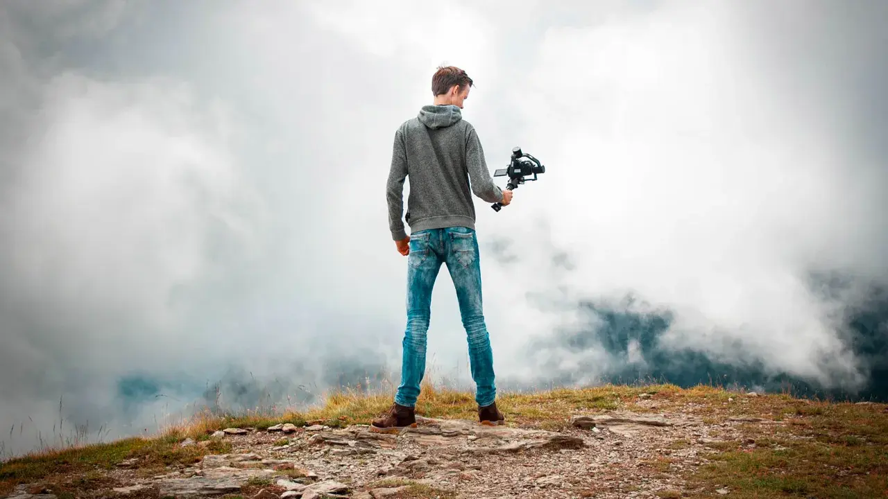 Joven en la cima de una montaña, sosteniendo un **gimbal** para estabilizar su cámara y capturar la vista entre las nubes.