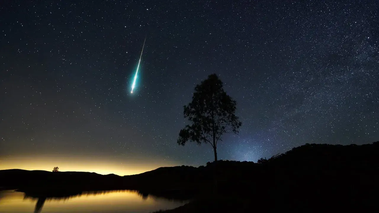 Un **obiettivo per fotografare le stelle** cattura una scia luminosa nel cielo notturno, con un albero solitario in silhouette e un lago calmo.