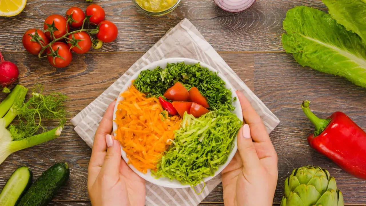 Manos sostienen un plato con una colorida ensalada mediterránea: zanahoria rallada, lechuga, perejil picado y tomates.