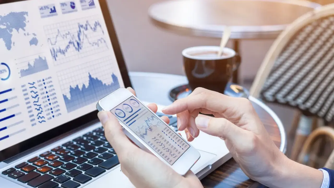 Manos revisando gráficos financieros en un teléfono móvil y portátil, gestionando el bbva net cash.