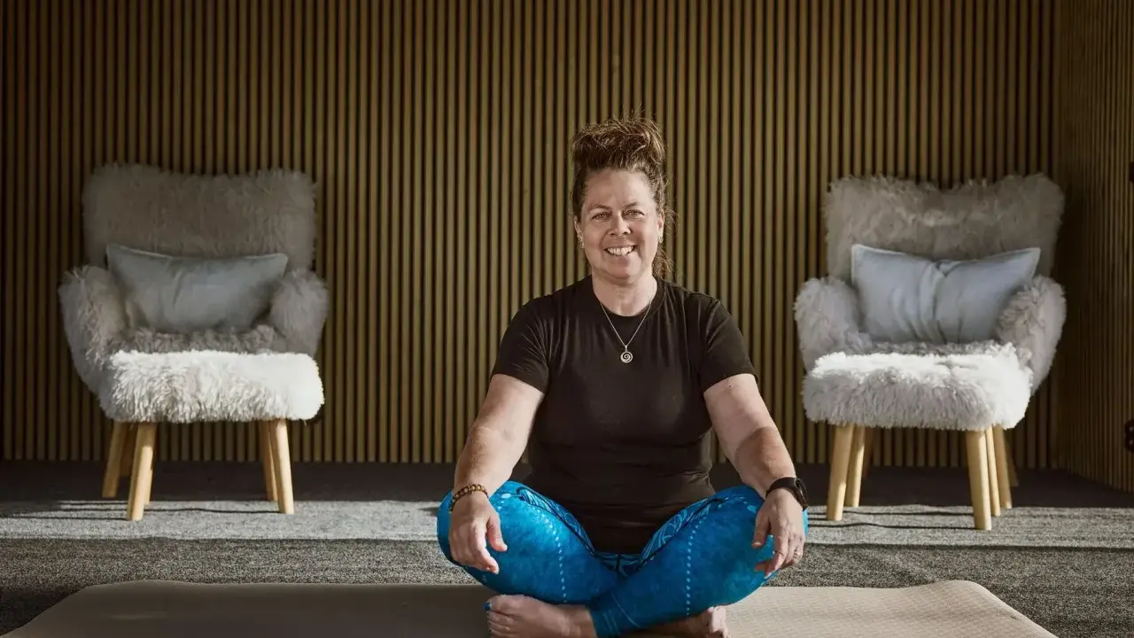 Kvinna i meditationsställning framför en vägg med träribbor. Två fluffiga fåtöljer står på var sin sida. Kanske hon funderar på hur hon ska dekorera hylla i sitt hem.
