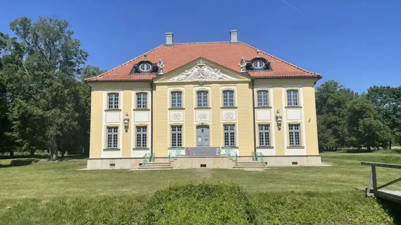 Piękny, żółty pałac w Choroszczy z czerwonym dachem, otoczony zielenią i błękitnym niebem.