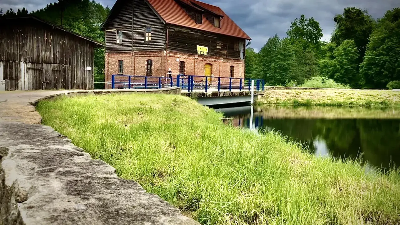 Stara ceglana budowla nad jeziorem Cekcyn, z drewnianą przybudówką i pomostem.