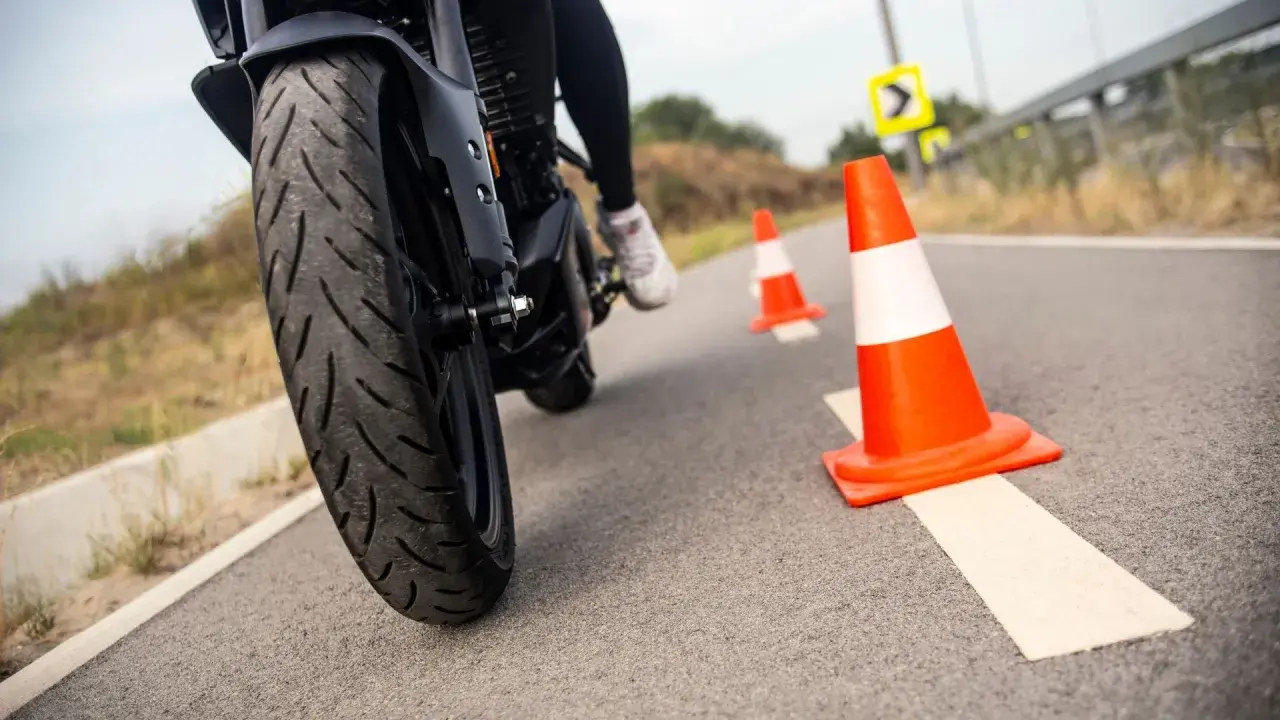 Motociclista practica maniobras entre conos naranjas. La rueda delantera de la moto, con su banda de rodadura, se ve de cerca.
