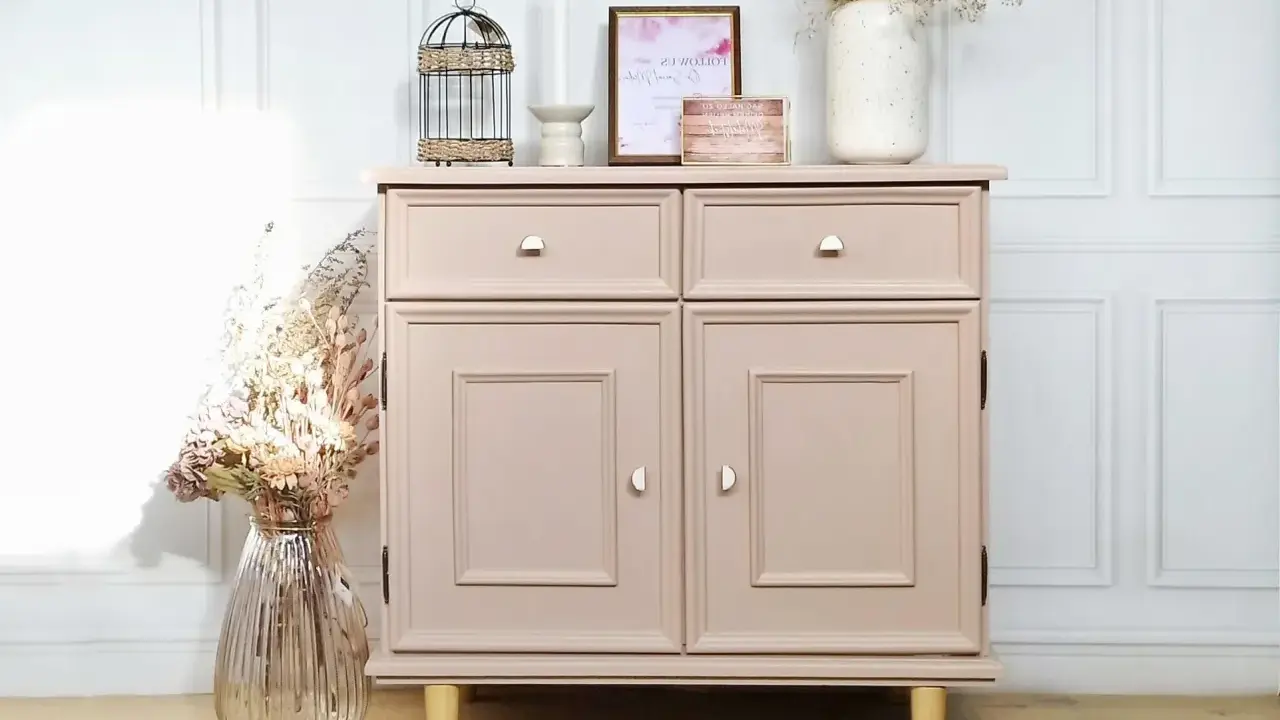 Meuble en bois peint en rose poudré, avec deux tiroirs et deux portes. Décoration florale et cage ornementale sur le dessus.