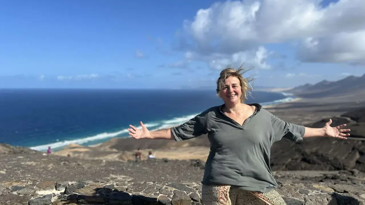 Donna sorridente con le braccia aperte su un paesaggio costiero, un'espressione di gioia che contrasta la solitudine interiore.